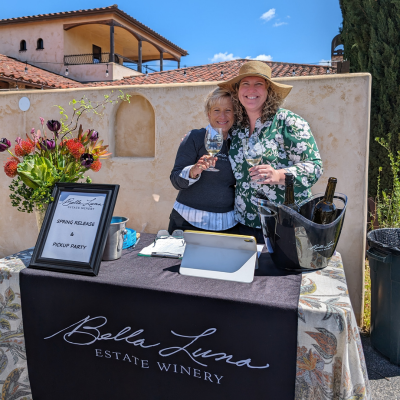 Two women smiling and holding wine glasses at Bella Luna Estate Winery tasting table in Paso Robles, showcasing a welcoming winery experience